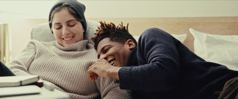 A woman wearing a sweater and grey beanie lies in bed next to a man wearing a blue sweater. Both are smiling, and the man is laying his head on the woman's arm.