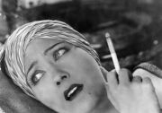 a close-up of a woman's face as she reclines. she wears a turban and smokes a cigarette. she looks worried.