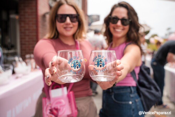 Two women holding up wine glasses with the "Rosé Day LA Wine Festival" logo. In the background, there’s a table with various items and other attendees, indicating an outdoor festival setting.