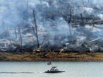 a small white speedboat with a prominent American flag mounted in the rear traverses in the foreground, as a forest is charred and burning in the background. 