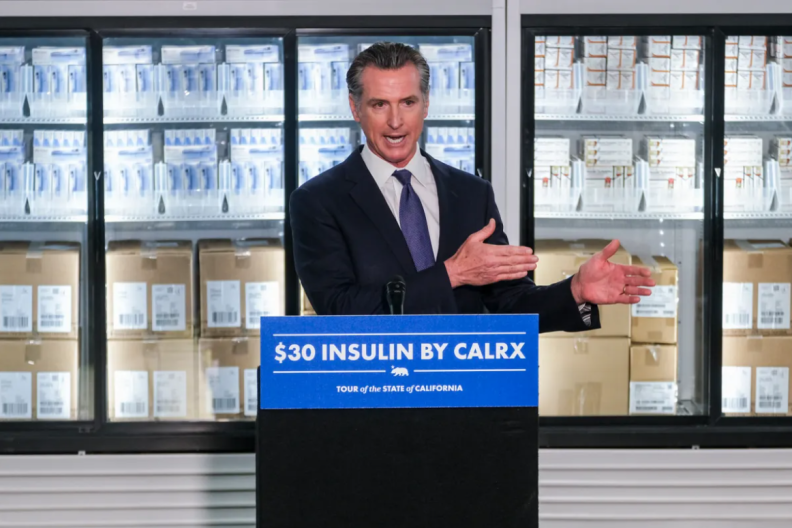 Governor Gavin Newsom stands in front of a podium with a sign that reads "$30 insulin by calrx" and behind him are a wall of fridges containing white and brown boxes.