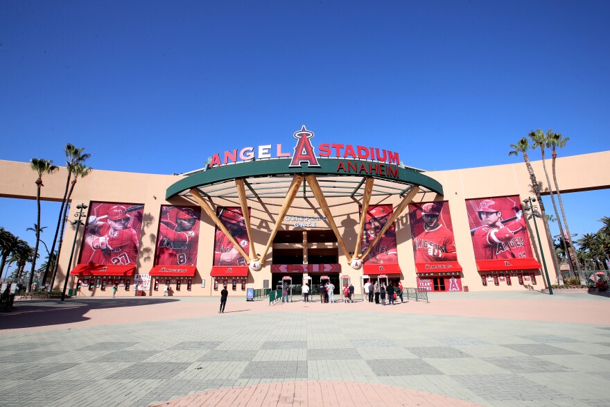 A massive tan marquee spans the entire background of the photo, with six large banners, about twenty feet tall, depicting different Los Angeles Angels baseball players wearing red uniforms. At center, an overhanging structure supporter by six large, tan baseball bats holds up a green railing that "Angel Stadium" with a large red "A" in the middle.