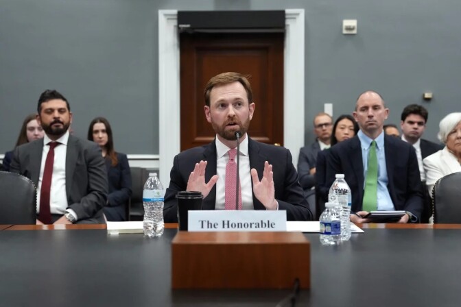 A man in a suit sits at a long table in front of a microphone. He is surrounded by water bottles and b.ehind him are men and women also in suits