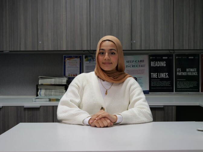 A brown skinned woman with a brown hijab and a white cardigan poses in a seated position in a room with one hand on top of the other. 