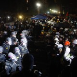 Several lines of police in helmets and other riot gear face off with protesters, many in hard hats and masks