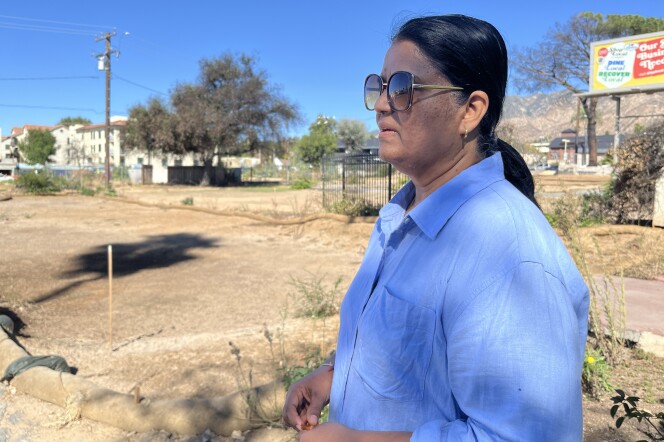 A woman with medium dark skin tone and blue shirt looks to the distance in front of an empty dirt lot.  