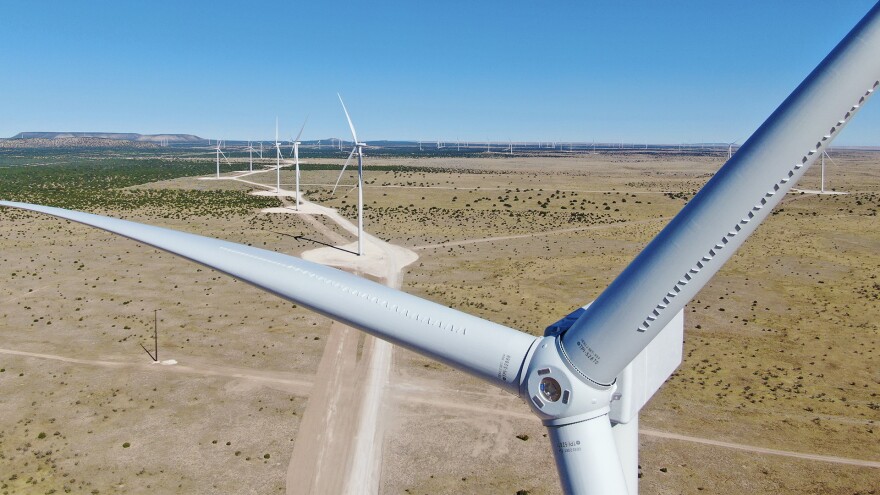 A white wind turbine in the foreground looms high above a desert landscape. White wind turbines span out into the distance connected by dirt roads.