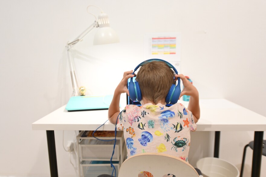 A student wearing a colorful shirt with sea animals on it and large over-the-ear headphones sits at a desk in front of a white wall.