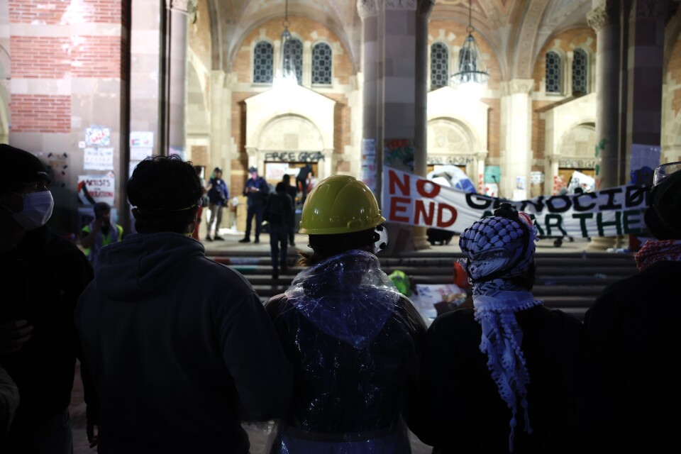 A row of people face a lighted building. A sign on a long piece of cloth reads: No Genocide