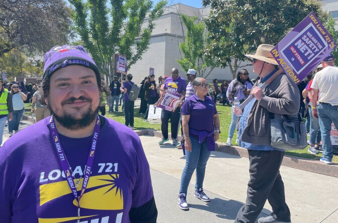 Man wearing purple hat and T-shirt smiles at a political rally. 