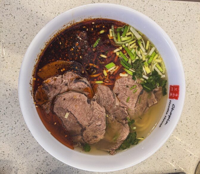 An overhead view of beef noodle soup in a white bowl printed with the Chopin Noodle House logo. Slices of beef sit in a clear broth with a pool of red chili oil on one side, topped with chopped leeks and cilantro, with noodles visible beneath.