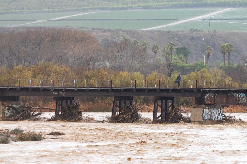 A tumultuous muddy river flows towards a bridge with train tracks. The stands of the bridge in the water are full of drift wood and debris.
