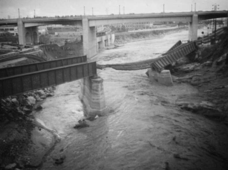  A vintage black-and-white image shows a part of a railway bridge, twisted and torn as if it weighed nothing heavier than paper. Beneath the wreckage runs the swollen LA River.