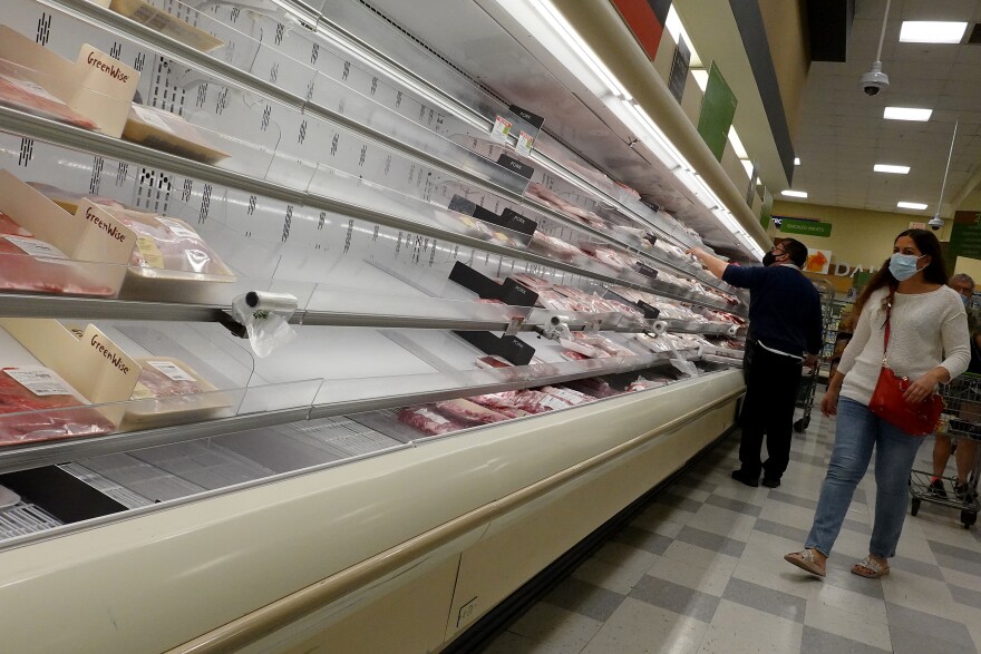 Empty refrigerated shelves in a grocery store where meat should be. Customers wearing masks walk by.