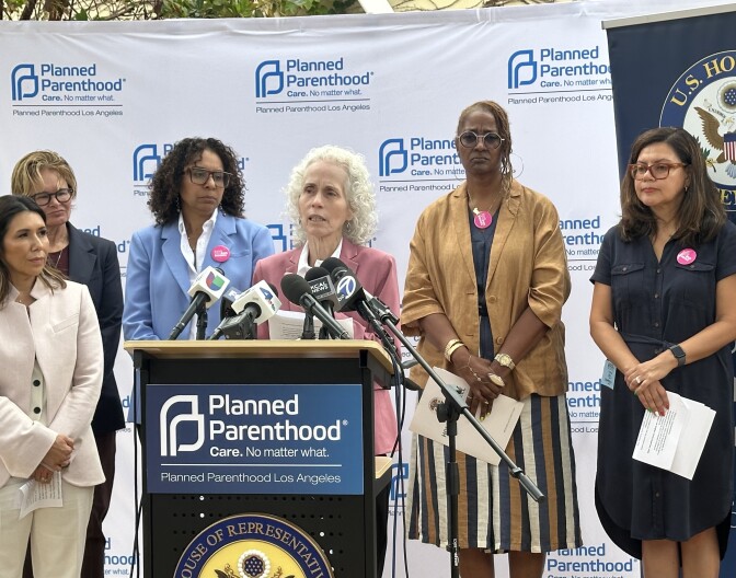 A group of women stands behind a podium at a Planned Parenthood Los Angeles press conference. The woman at the podium, with curly white hair and a pink blazer, speaks into microphones from multiple news outlets including Univision, NBC 4, and ABC 7. Surrounding her are six women, some wearing “Bans Off Our Bodies” buttons. The backdrop features Planned Parenthood logos, and a U.S. House of Representatives seal is partially visible on the right.