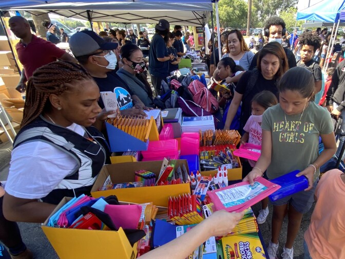 Children and their parents line up in a parking lot under a canopy at outdoor tables to collect in a variety of donated school supplies. Volunteers are on the other side of the tables handing out the supplies.