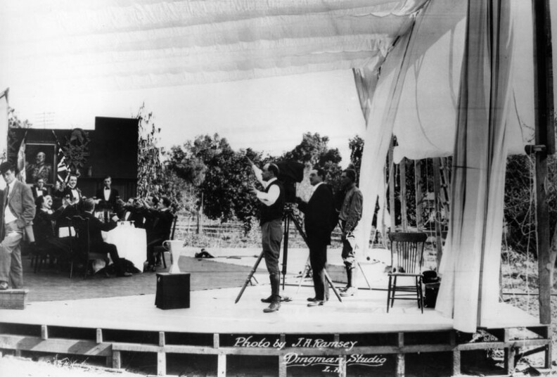 The filming of the first scene of "The Squaw Man" at Lasky Feature Play Company, which later combined with the Famous Players Co., later the Paramount Famous Lasky Corporation. Cecil B. DeMille, director of the film, is in the center of the picture, wearing boots and a light suit. Dustin Farnum, star of the production, is on the stage, his hand raised, while the camera grinds. Cameraman is Alfredo Gandolfi. The building on the left is the old Lasky Barn, once situated in a lemon and orange grove on Vine Street and Selma Avenue. The barn occupied a place of honor on the new studio lot and is now open to the public as the Hollywood Heritage Museum on Highland Avenue. 