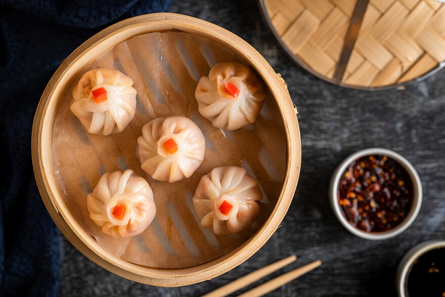 Five glistening dumplings sit inside a bamboo steamer, a pair of chopsticks at the ready, alongside of bowl of garlic-studded chili sauce.