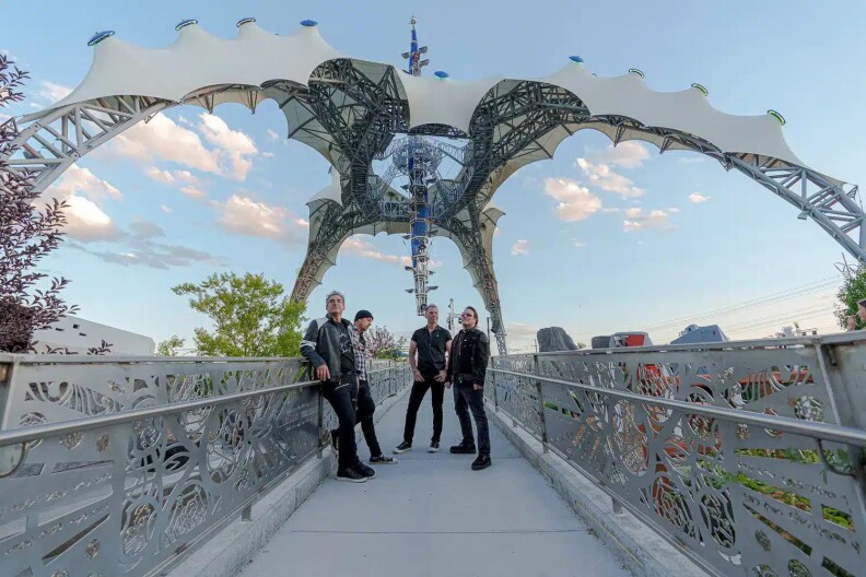 The four members of the Hollywood U2 cover band post together on a bridge during the daytime. They're all wearing black shirts or jackets, with one person wearing a checkered black and white button-down.