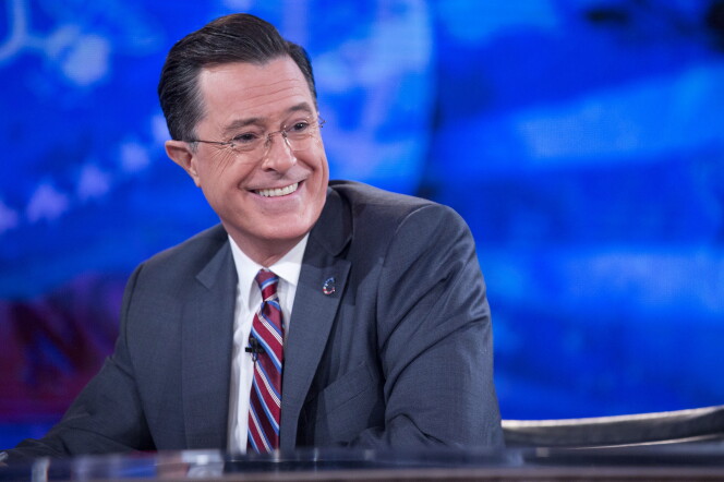 Stephan Colbert, a man with light light skin tone wearing a gray suit, striped tie and glasses, smiles as he looks out of frame to his left.