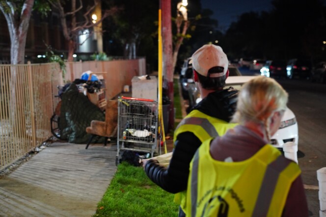 Two people wearing reflective vests stand next to a makeshift shelter on the sidewalk.