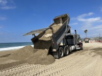 A truck spills a load of sand onto the beach on a day bright with sunshine. Orange work cones and another truck are visible in the background, underscoring the massive job underway.  