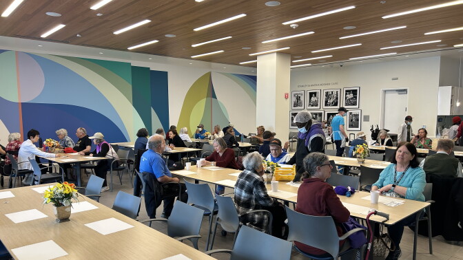 A senior center room filled with at least 7 dining tables with 8 seats each. Older adults are sitting across the tables, some with lunch trays and paper cups in front of them. At least two men can be seen walking the room in aprons and hairnets, handing out food to the older adults.