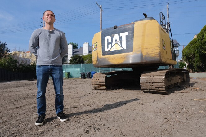 A man with light-tone skin stands with his hands behind his back in a dirt lot. A CAT bulldozer is behind him. 