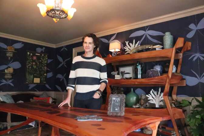 A man with fair skin who appears to be in his 30s stands behind a wood dining room table, smiling. Behind him is a large wood bookcase with art objects, a lamp and plants. 