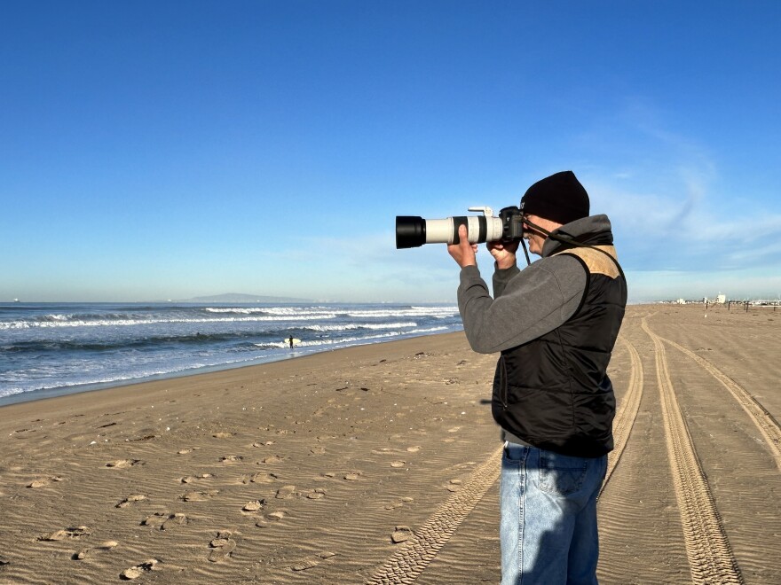 A white man wearing a black beanie, jeans and sweatshirt stands on a sunny, wide beach pointing a camera with a long lens at the ocean.