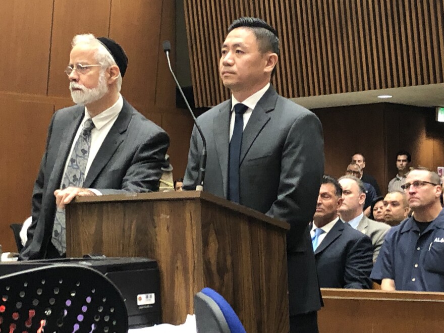 Luke Liu at his Dec. 11, 2018 arraignment. He stands in a gray suit with a black tie and white shirt at a wooden podium with a thin microphone snaking into the air in front of him. He is to the right, and to the left is his attorney Michael Schwartz, wearing a grey suit and tie and a white shirt. He has glasses and a full beard and mustache, and is wearing a yarmulke. Several people in the audience in the courtroom can be seen seated behind them.