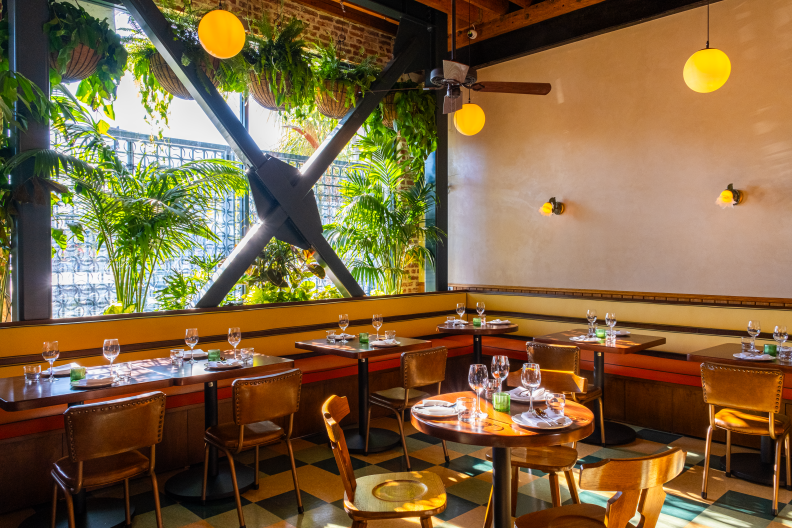 The restaurant's interior features a spacious corner with yellow banquettes, wooden tables, and leather-bound chairs where two walls intersect. One wall includes a large window secured with a grey metal grate, partially concealed by a plant that overlooks the outside. The opposite wall is painted off-white and is illuminated by two sconces.