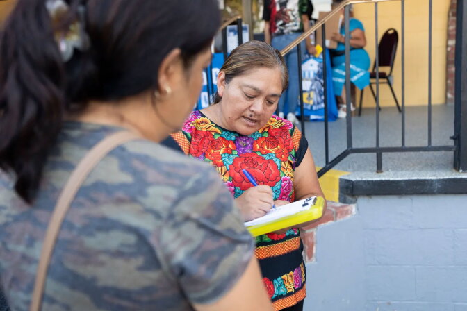 A person in an embroidered floral top writes on a clipboard while speaking with someone at an outdoor event.