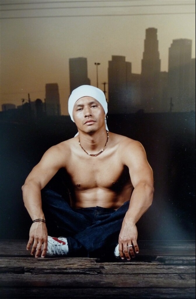 A brown-skinned man sits with his legs crossed in a professional photo. 