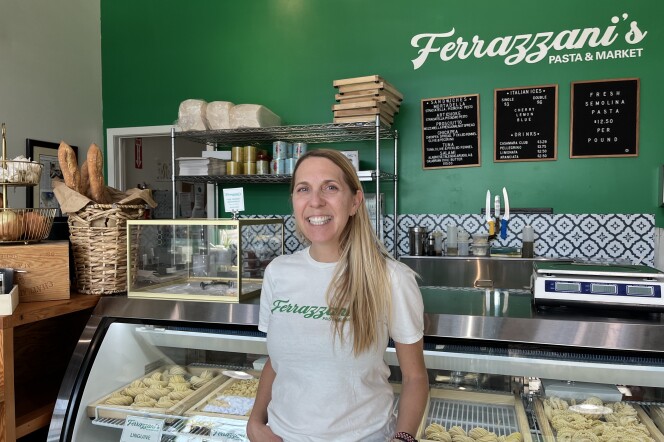 A light-skinned woman stands in front a stainless steel counter with a glass display and trays of rolled pasta behind it. The woman is wearing a white t-shirt. The green wall behind her has a black menu board and a white lettering that reads "Ferrazzani's Pasta & Market."