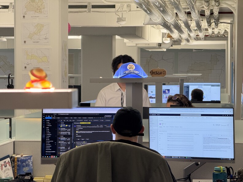 A man sits in an office chair facing two computer screens and a shelf with a blue hat.