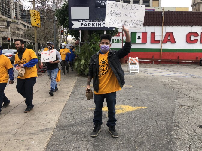 A man in a yellow shirt and black jacket stands in a parking lot beside a sidewalk holding a sign in one hand that reads "Gov. Newsom Save our solar jobs." 