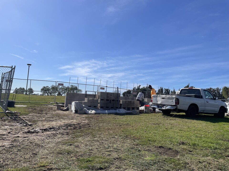 A wide show showing two men among a pile of cement blocks next to a white pickup truck. There's fencing in the back. It's a blue-sky day in a grassy and muddy field. 