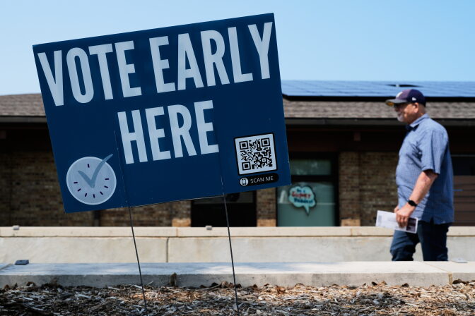 A person, slightly out of focus in the background, walks behind a sign in the foreground that reads "Vote Early Here."