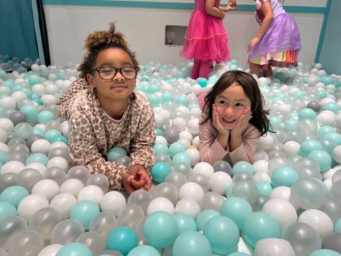 A female with medium skin tone and brown, curly hair and another female hair with light skin tone and brown, wavy hair sit in a ball pit with turquoise, grey and white balls. 