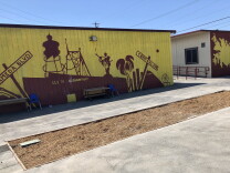 A rectangular patch of dirt is in front of a building with a yellow and magenta mural. The mural depicts Watts landmarks such as the Watts Towers and street signs for Central Avenue and Alameda Boulevard.