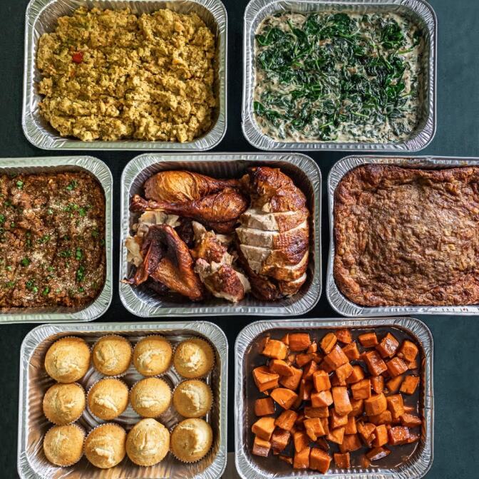 Overhead photo of large aluminum catering trays filled with Thanksgiving dishes, including smoked sliced turkey, cornbread muffins, candied yams, creamed spinach, cornbread dressing, mac and cheese, and a baked casserole.