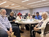 Six men and women, all older adults, are sitting around a white plastic table in a large interior room. White trays of food are placed in front of each of their seats.