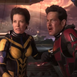 Evangeline Lilly and Paul Rudd stand together in a futuristic space ship. Lilly wears a black and yellow suit that is decoratively segmented into metal armored plates. The suit covers her whole body, only her head visible. Her mouth is slightly opened and brows slightly furrowed looking at someone out of frame. She grasps the right arm of Rudd with her left hand. Rudd wears a similar armored suit but in red and black. His head is also visible. Blood streaks down his face from his nose. He looks flushed with his mouth open and eyebrows pushed together. 