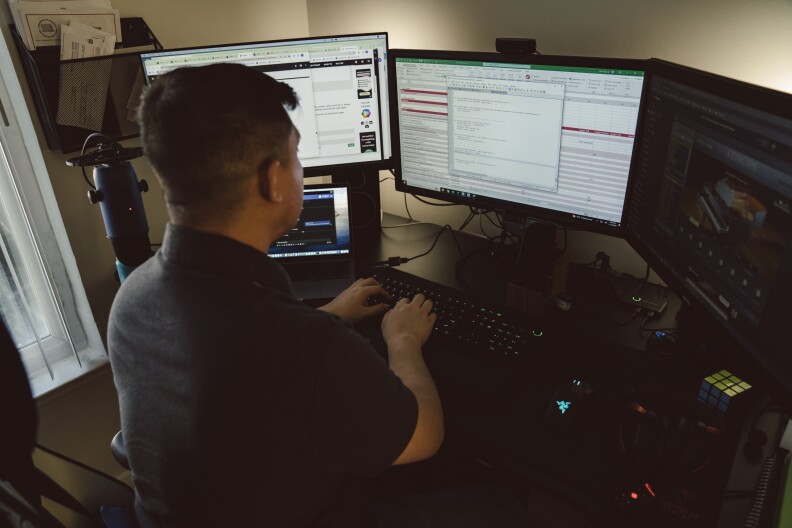 A Filipion man wearing a Black shirt, sits at a chair, his back faces the camera and he faces three computer screens while typing.