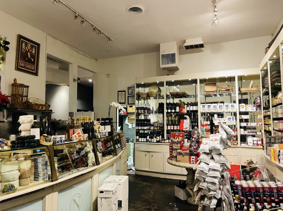 The interior of a store packed with bottles of wine, packets of crackers and other delicious looking things for sale. Behind a glass counter are segments of different cheeses