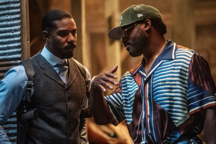 Two dark-skinned men are facing each other. One is dressed in a suit and the other is wearing a baseball cap and collared shirt.