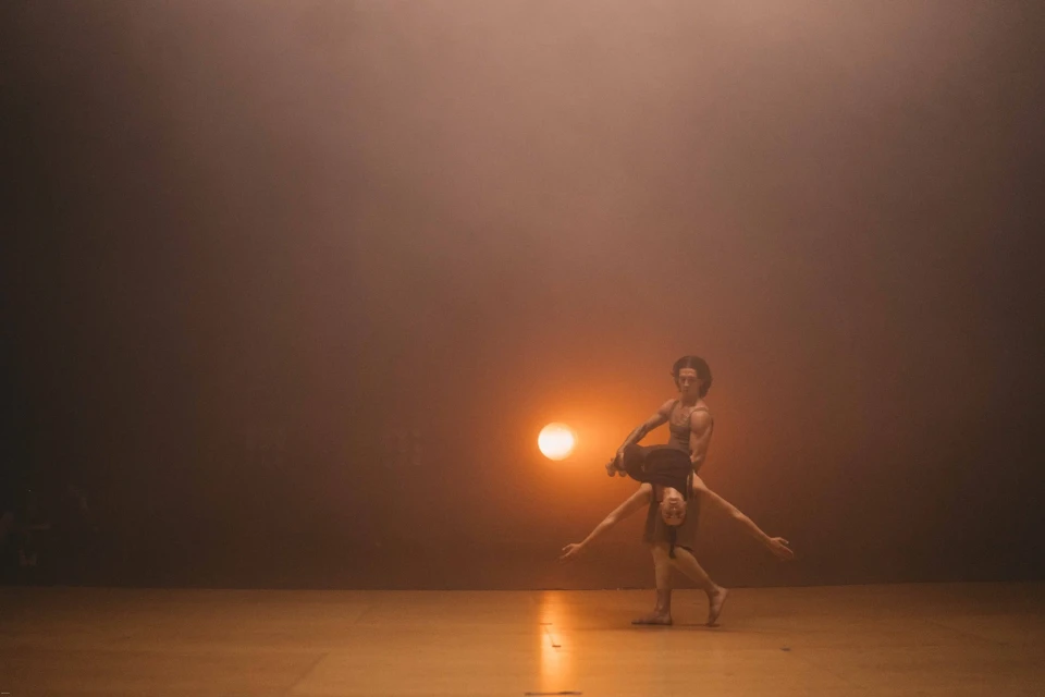 A man holds a woman sideways with her arms out on a dance stage with a single spotlight behind them. 