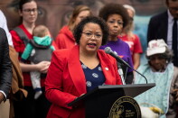A woman wearing a red blazer and blue and white blouse stands at a podium, speaking into a microphone. A group of people stand behind her.