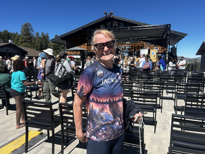 A smiling person wearing sunglasses and a nature-print shirt reading 'Jackie' stands near rows of chairs and an outdoor stage.